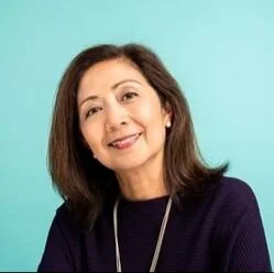 Portrait of a woman with dark brown hair wearing a navy blue top and a necklace against a teal background.