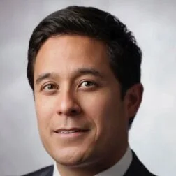A headshot of a man with dark hair, wearing a dark suit and tie, smiling calmly against a plain gray background.