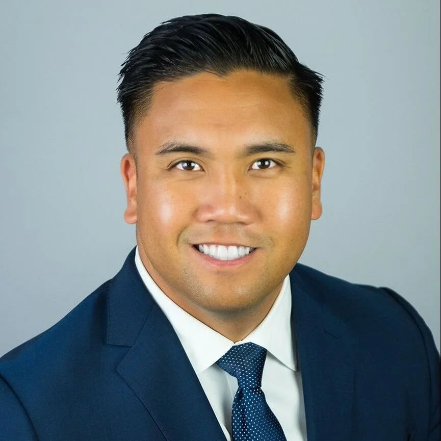 A professional headshot of a smiling man with dark hair, wearing a navy blue suit, white shirt, and dark blue tie, against a plain gray background.