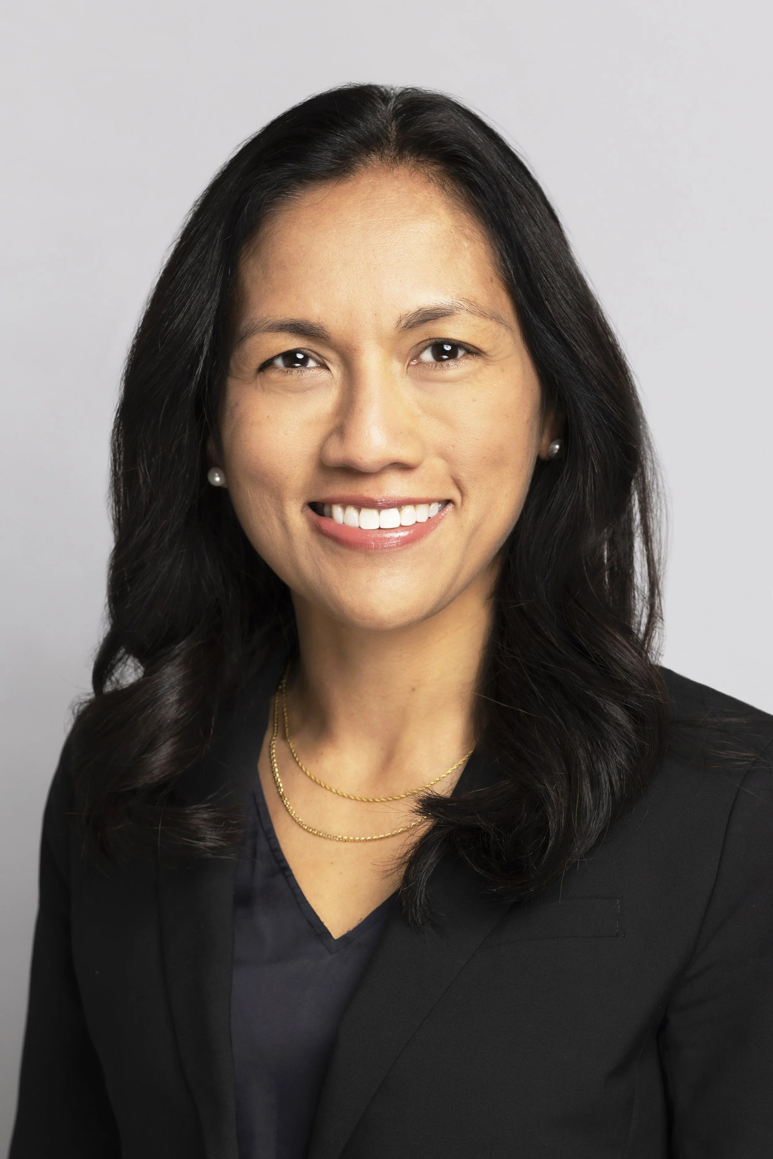 Professional headshot of a woman with long dark hair, wearing a black blazer and jewelry, smiling against a plain gray background.