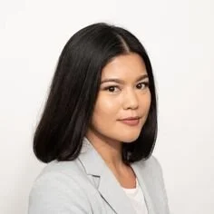 A woman with shoulder-length dark hair, wearing a light gray blazer, looking at the camera against a plain white background.