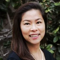 Portrait of a woman with dark hair smiling outdoors against greenery background.