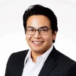 Professional headshot of a smiling man with glasses wearing a black suit and white shirt against a white background.