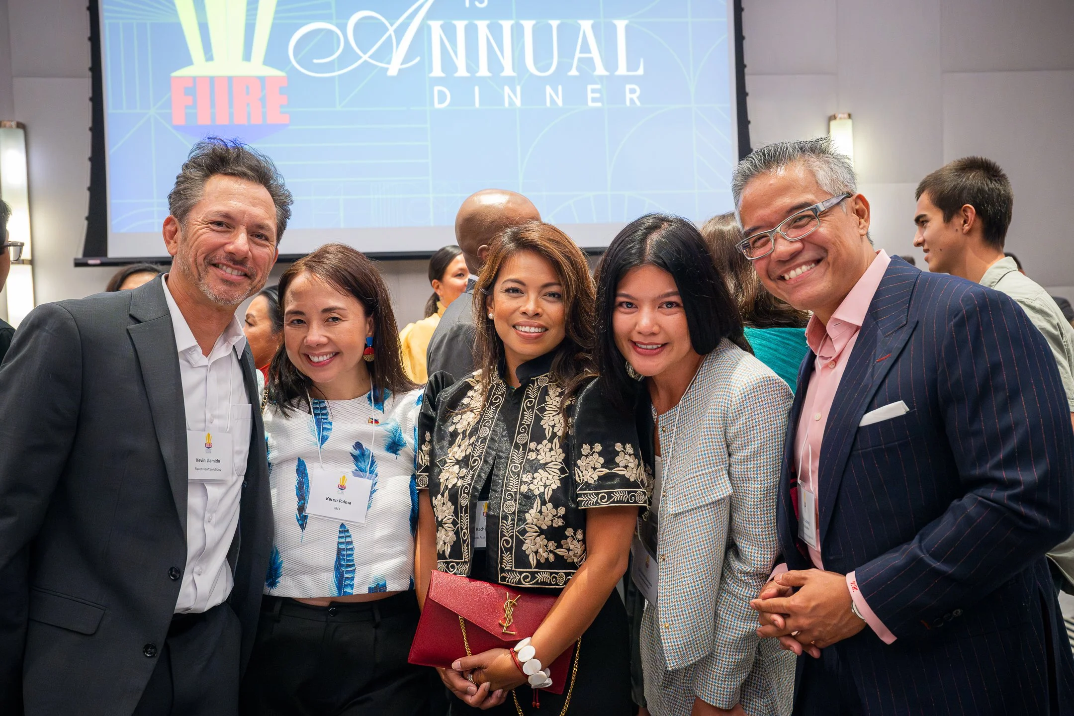 Group of five diverse people smiling at an annual dinner event, with a large screen in the background displaying the event's name.