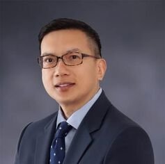 Professional headshot of a man in a suit with glasses against a gray background.