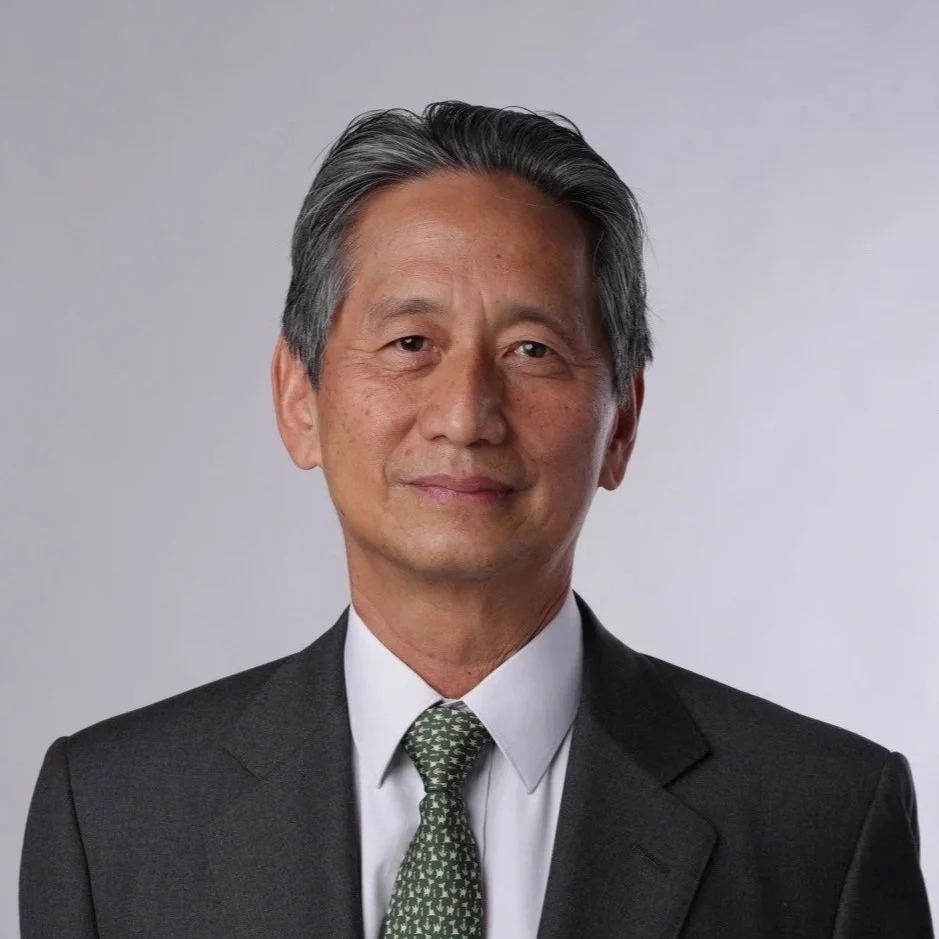 A middle-aged Asian man with graying hair dressed in a dark gray suit, white shirt, and patterned green tie, standing against a plain light gray background.