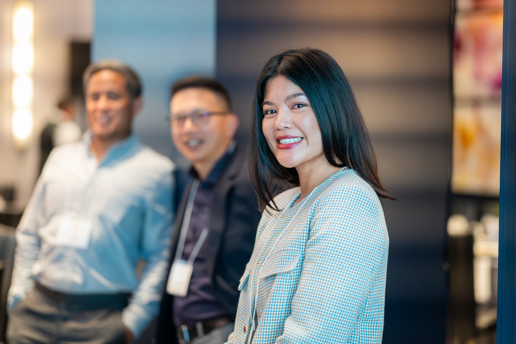 Smiling woman in yellow checked blazer with two men in the background at a professional event.