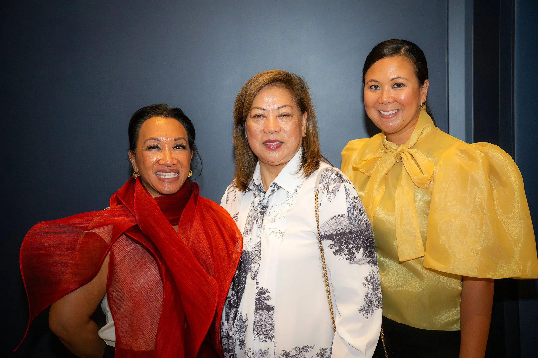 Three women standing together and smiling in front of a dark background, one wearing a red blouse with a large red scarf, one wearing a white blouse with black and white print, and one wearing a yellow blouse with puffed sleeves and a bow at the neck