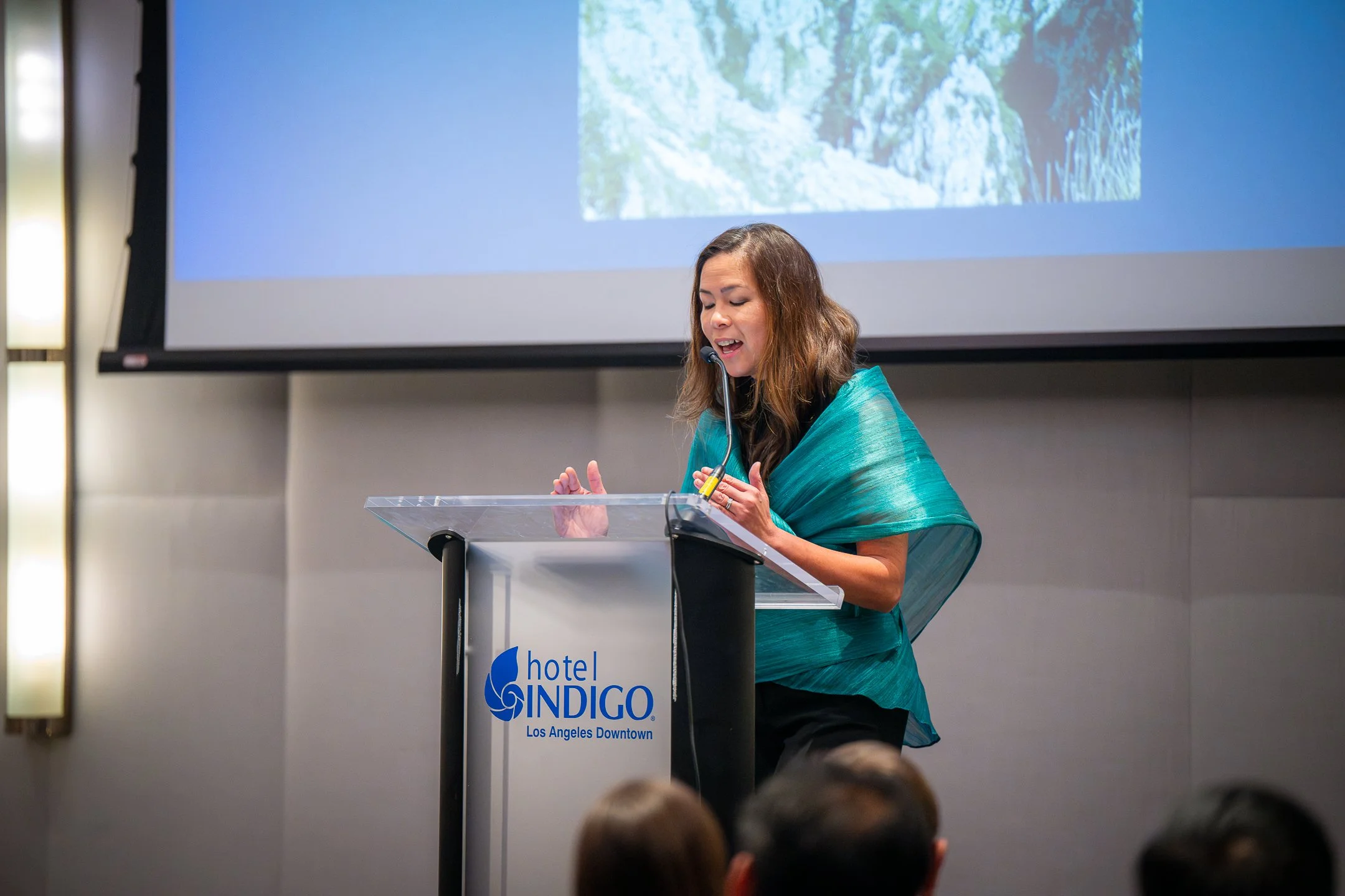 A woman in a blue and green shawl speaking at a podium with a microphone during a presentation at Hotel Indigo in Downtown Los Angeles.