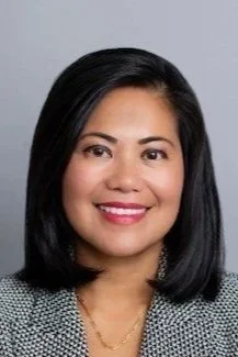 Headshot of a woman with shoulder-length black hair, smiling, wearing a light-colored blazer with a necklace, against a neutral gray background.