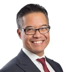 A man with glasses and dark hair, dressed in a suit and tie, smiling against a white background.