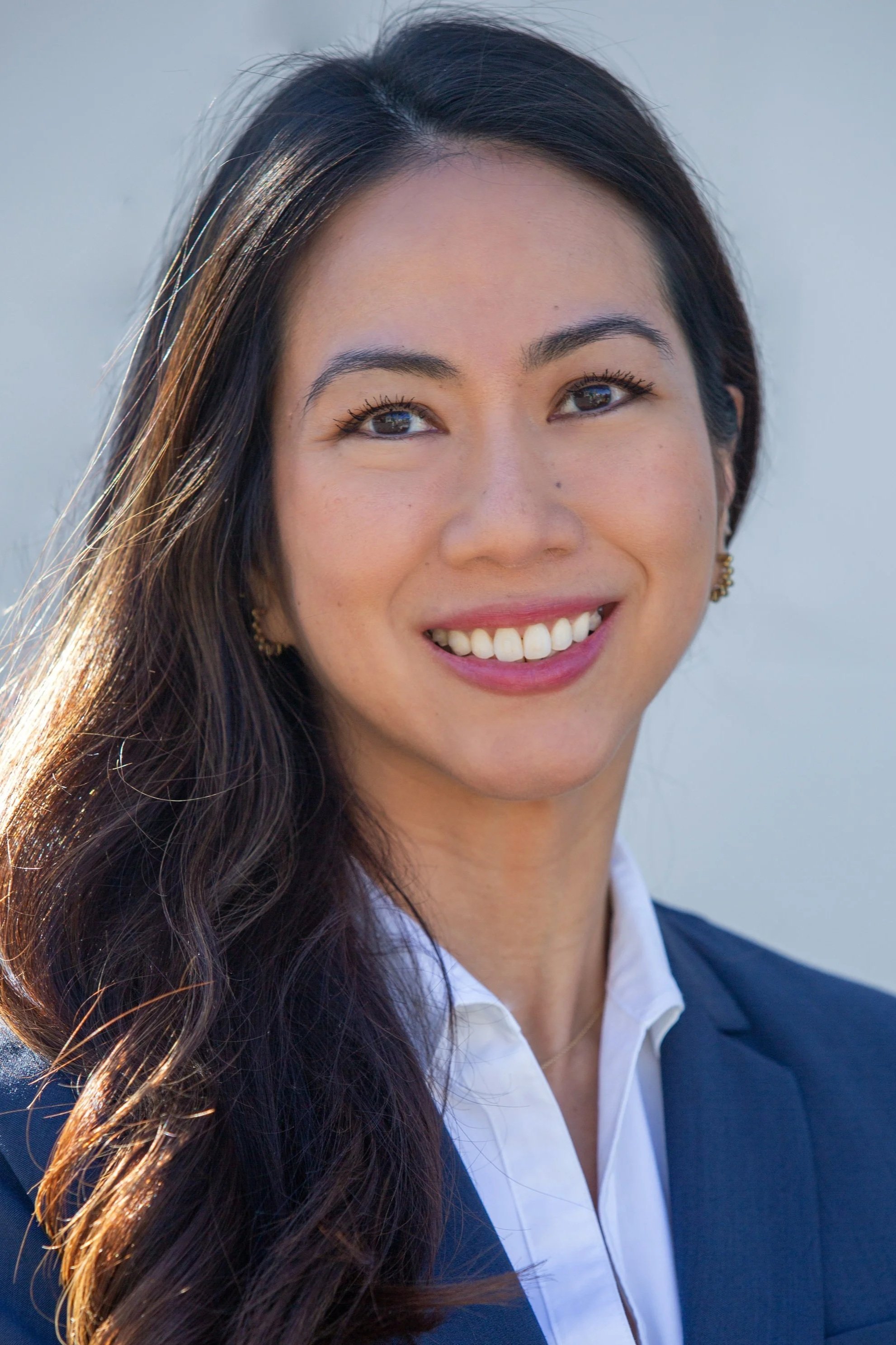 Headshot of a woman with long dark hair, smiling, dressed in a business suit, outdoors with natural light