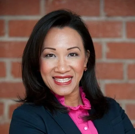 A woman with shoulder-length black hair smiling, wearing a navy blazer over a pink blouse, standing in front of a brick wall.