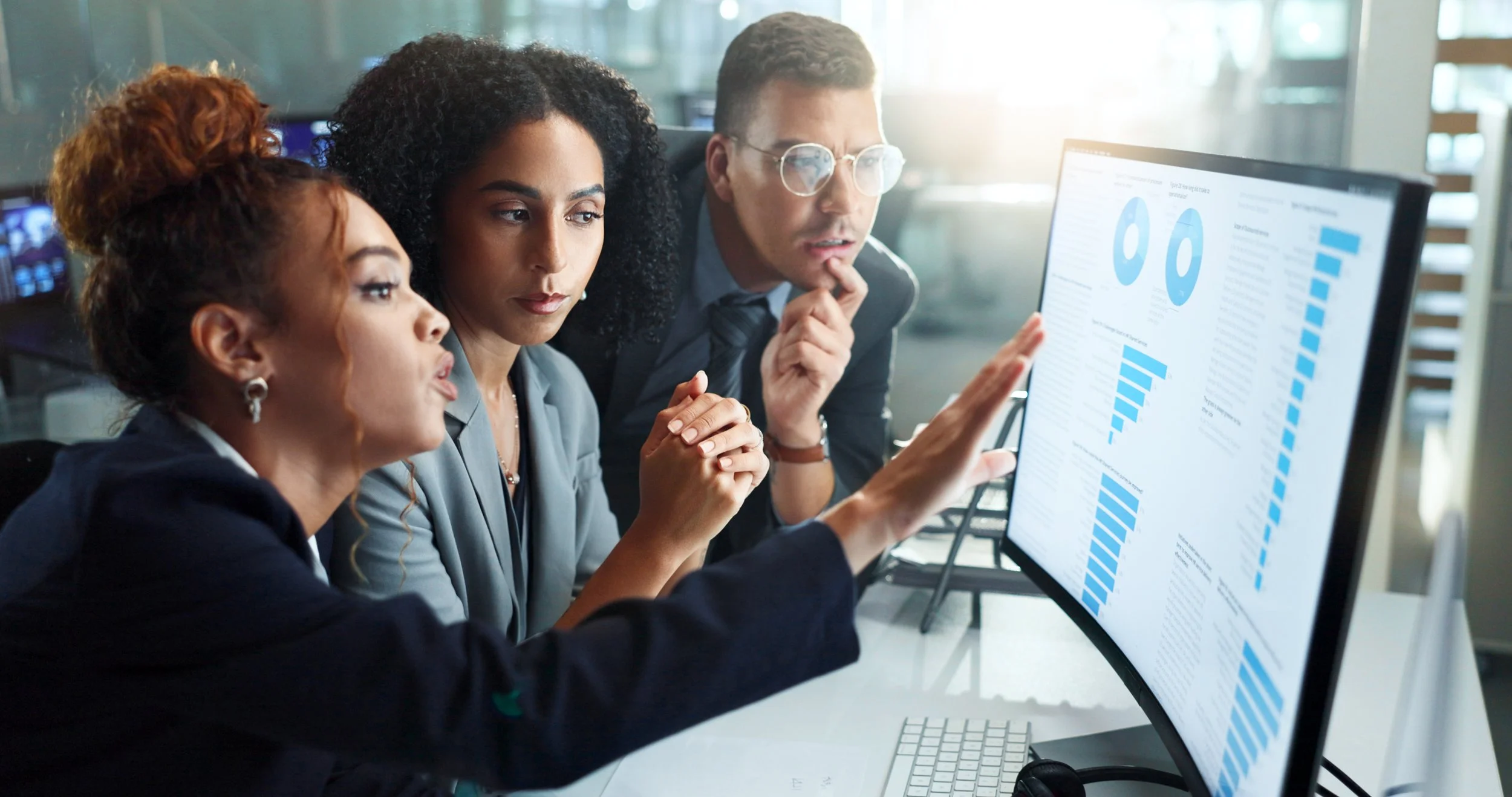 Three professionals in business attire analyzing data charts on a large computer monitor in a modern office.
