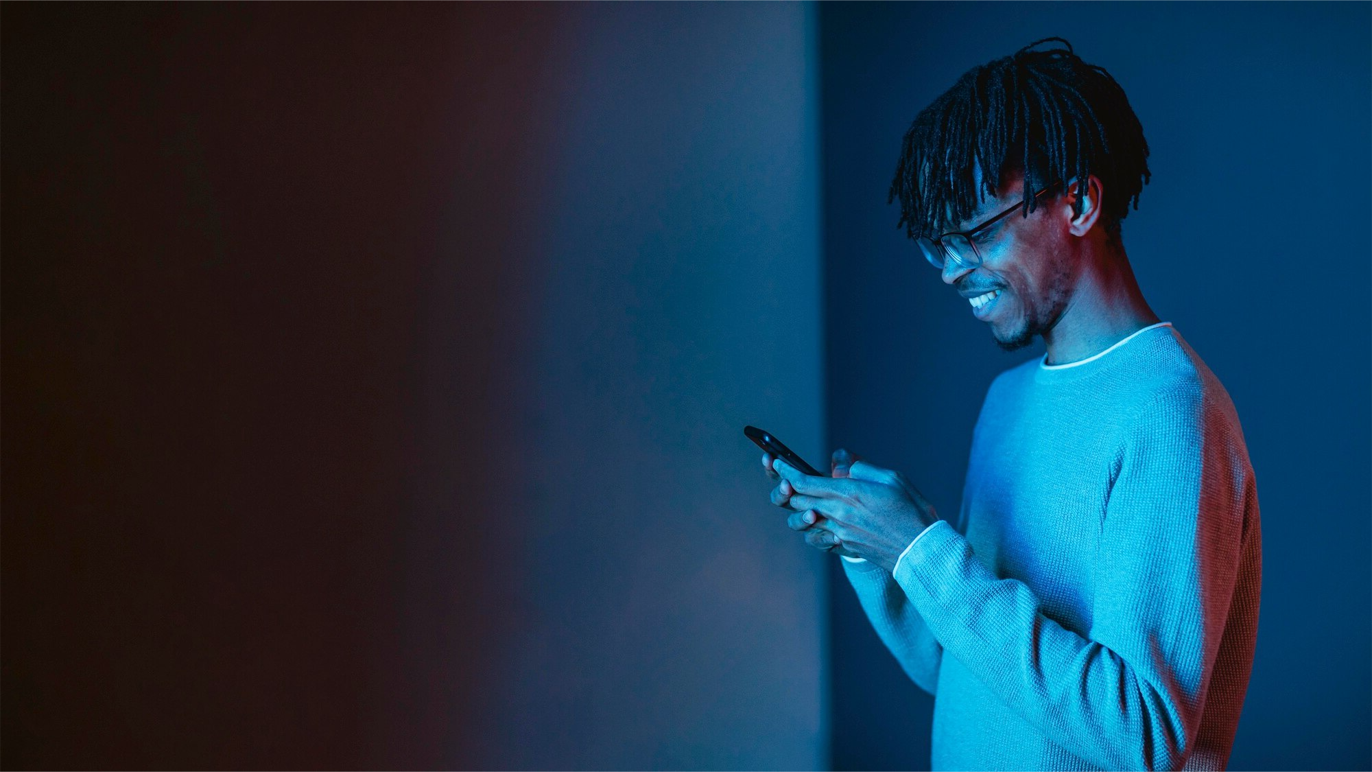 A young man with glasses and dreadlocks smiling while looking at his smartphone in a dimly lit room.