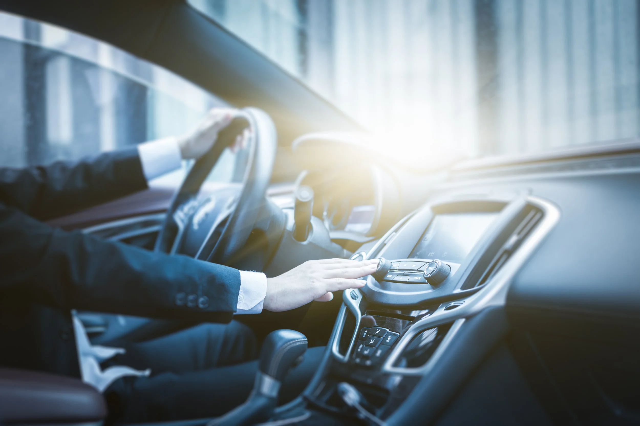 Person in business attire operating a car's dashboard and steering wheel during daytime with sunlight outside.