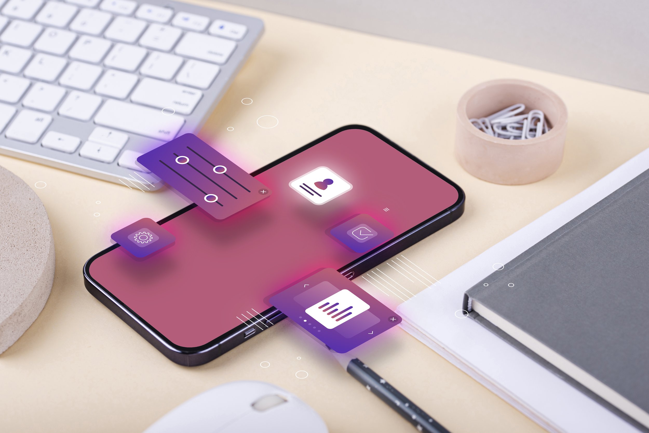 A smartphone displaying a digital interface with icons, placed on a desk with a white keyboard, a small pink container with paperclips, a gray notebook, a pencil, and a computer mouse.