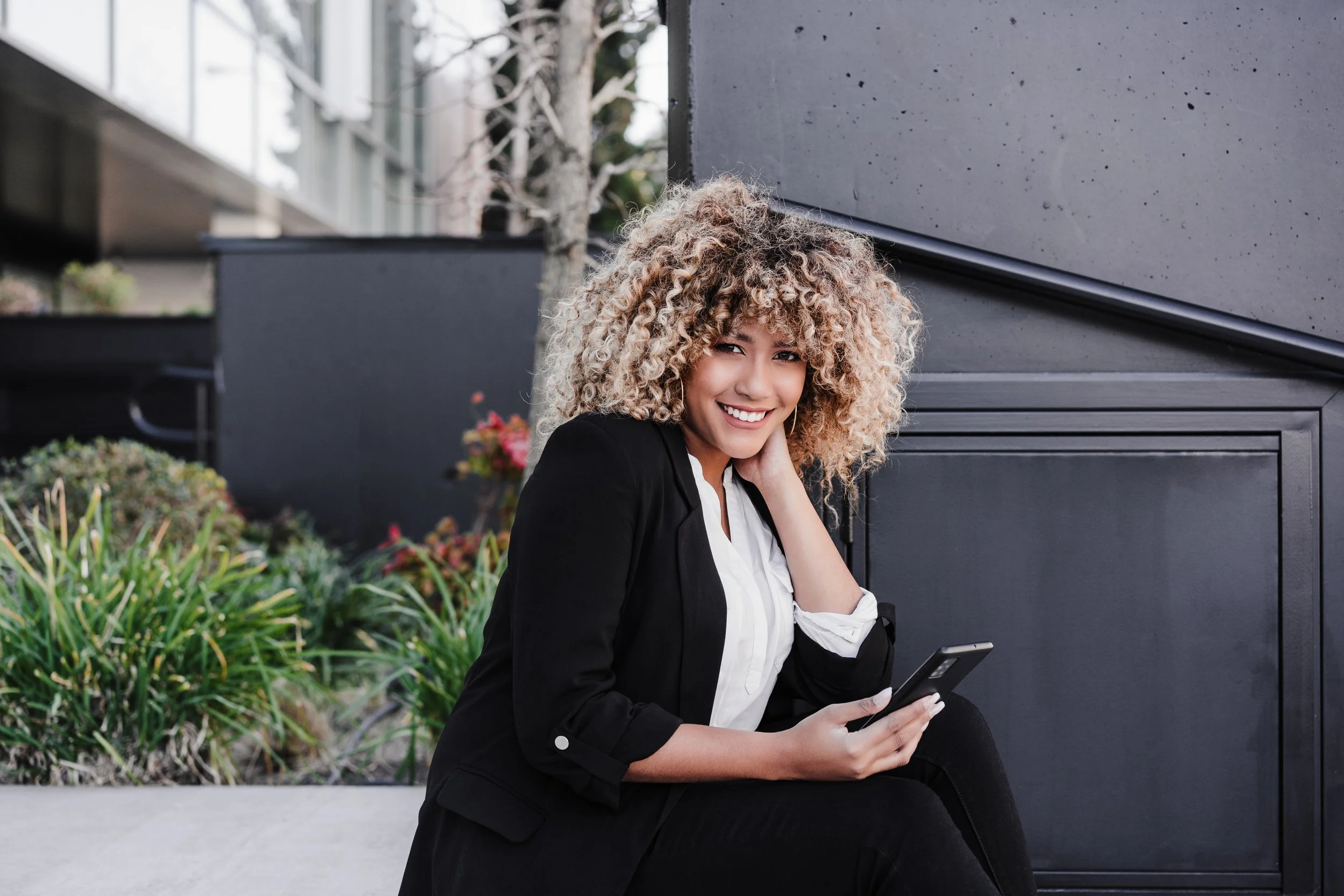 Young woman with curly blonde hair smiling and holding a smartphone outdoors, sitting on a concrete ledge near plants and modern buildings.