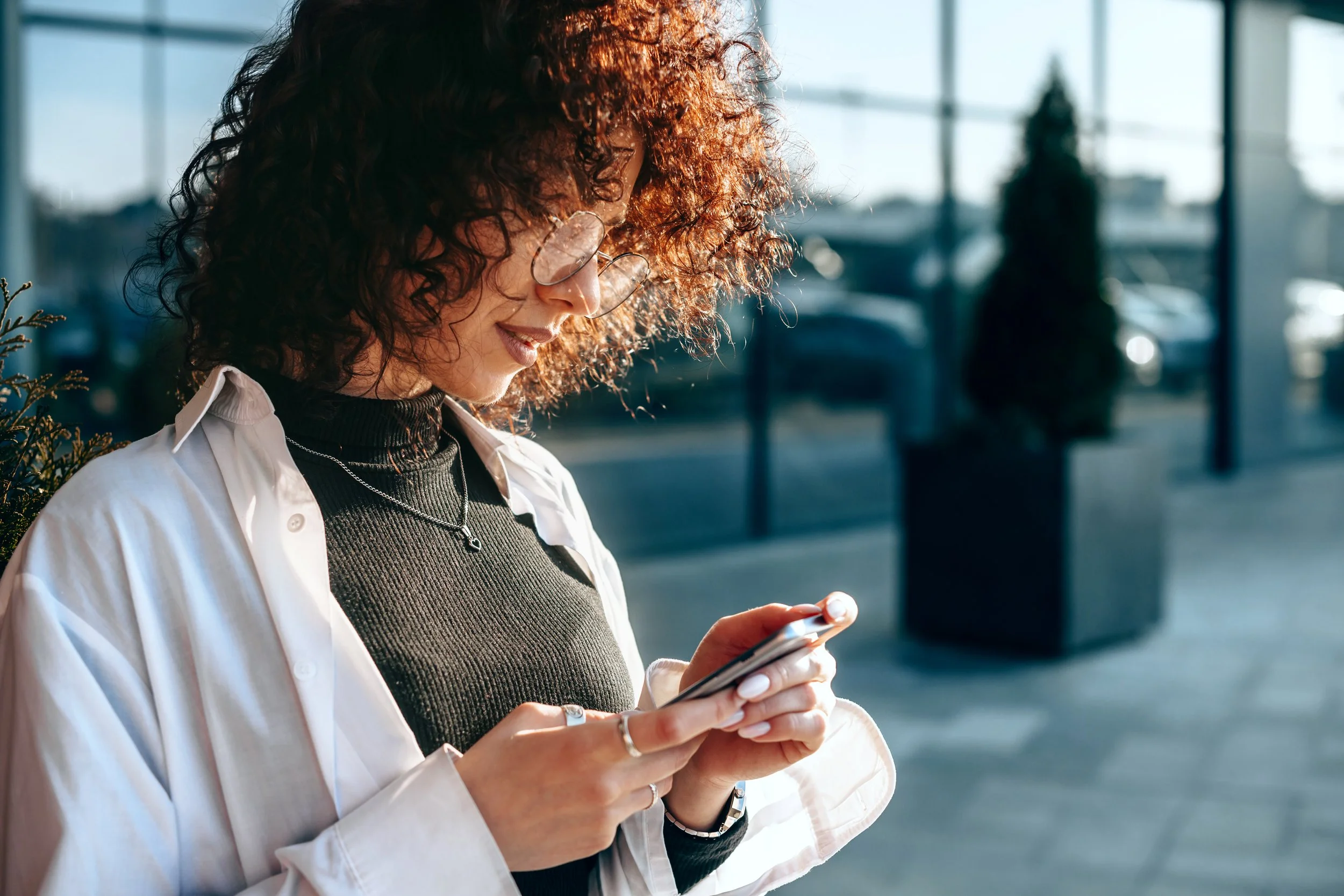 A woman with curly hair, glasses, and jewelry looking at her smartphone outdoors near a window with reflections of cars and trees.