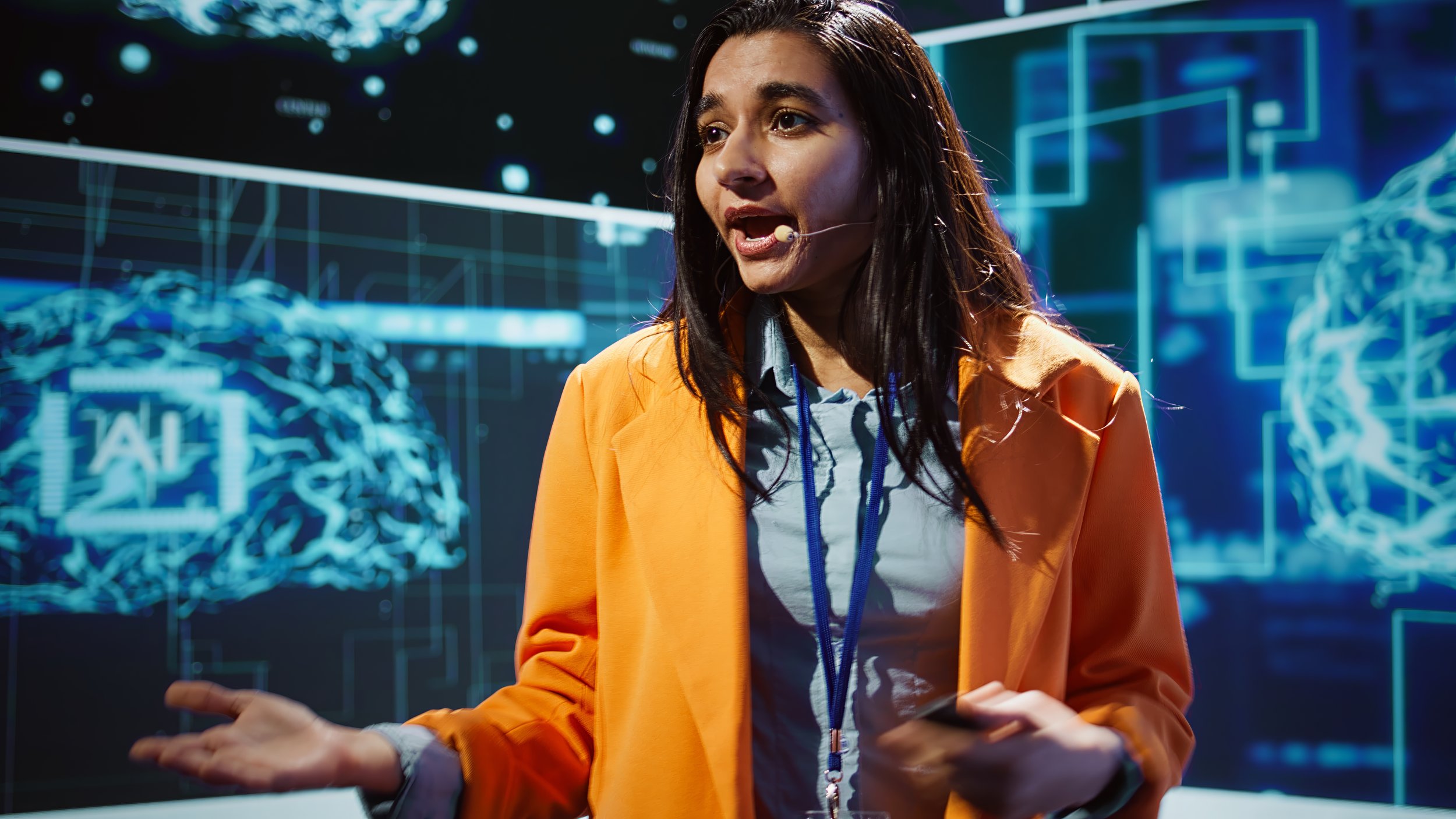 A woman in an orange jacket and headset is speaking in front of a digital display with holographic images of brains and technical graphics.