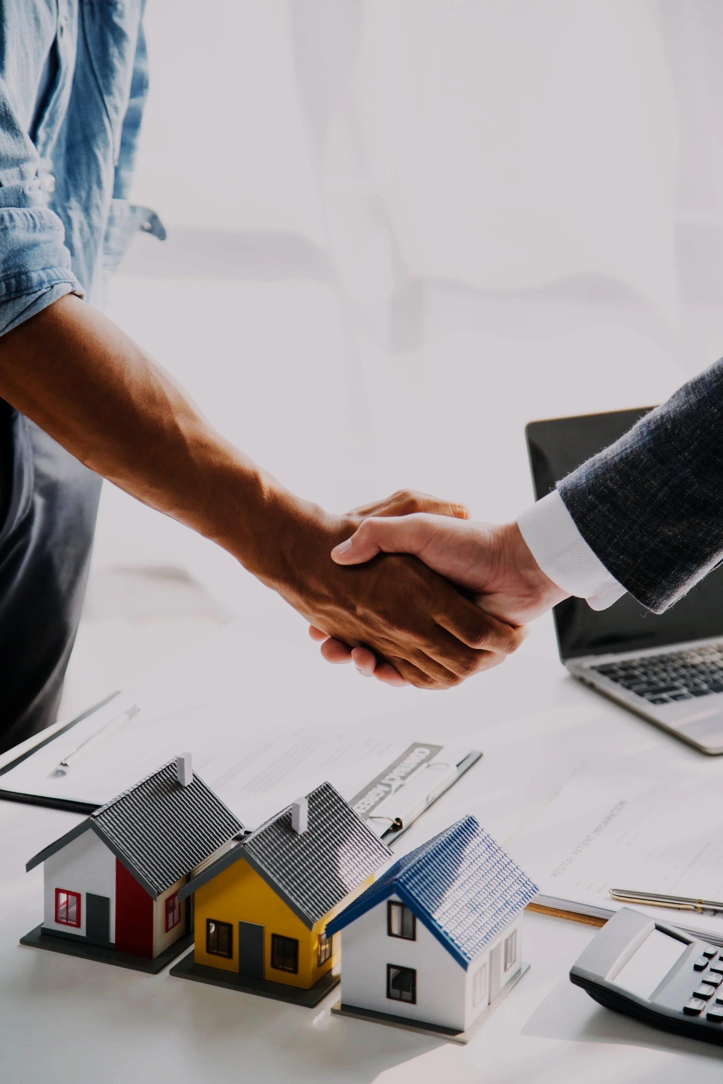 Two people shaking hands over a table with miniature house models, documents, a pen, a clipboard, a calculator, and a laptop.
