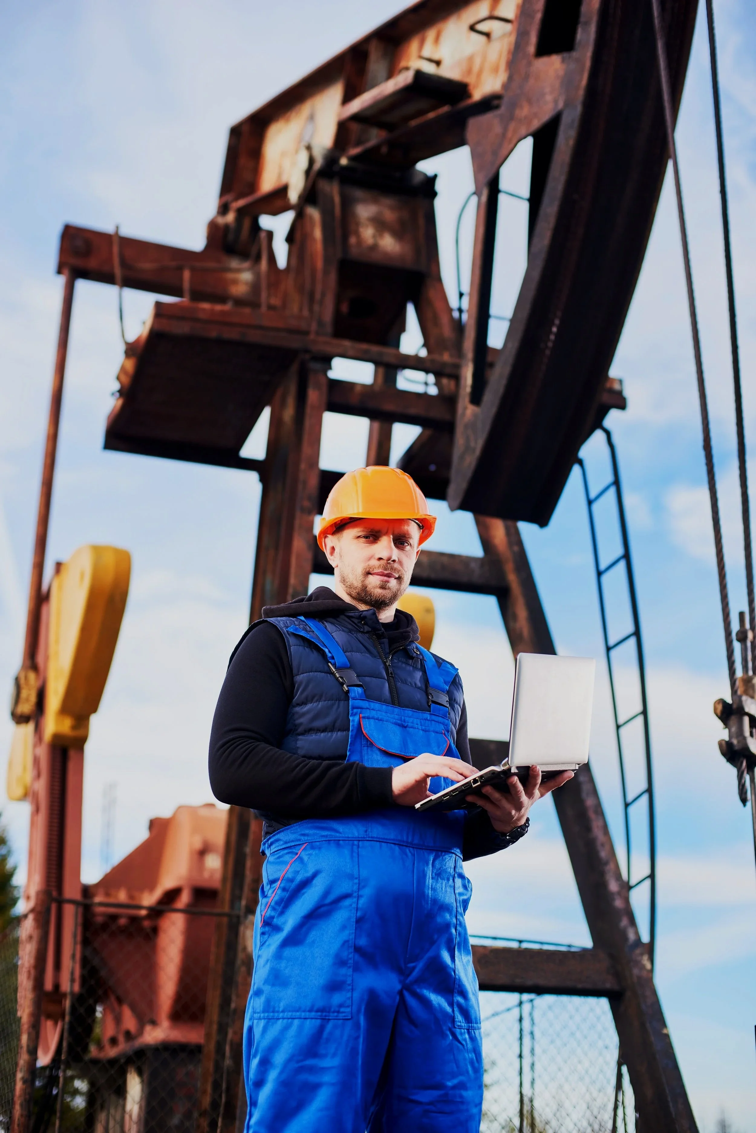 A man in safety gear, including a yellow helmet and blue coveralls, holding a laptop outdoors near a rusted oil derrick.