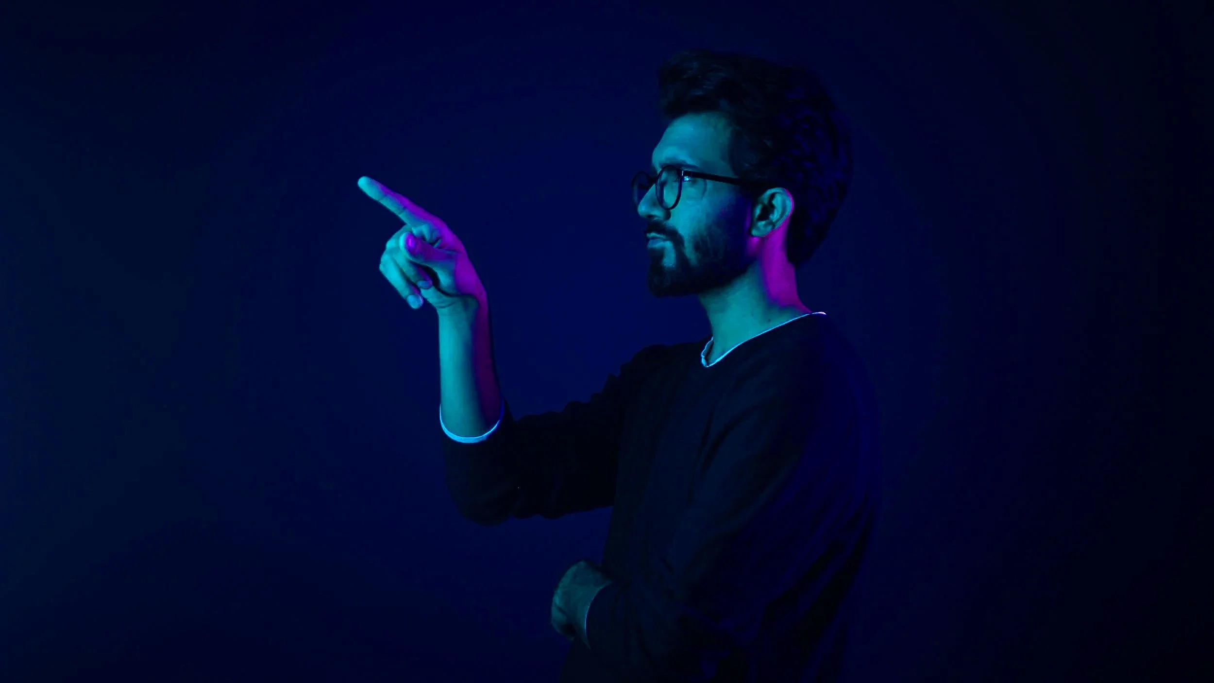 A man with glasses and a beard is standing in a dark space with a black background, illuminated by blue and purple lighting. He is pointing to the left.