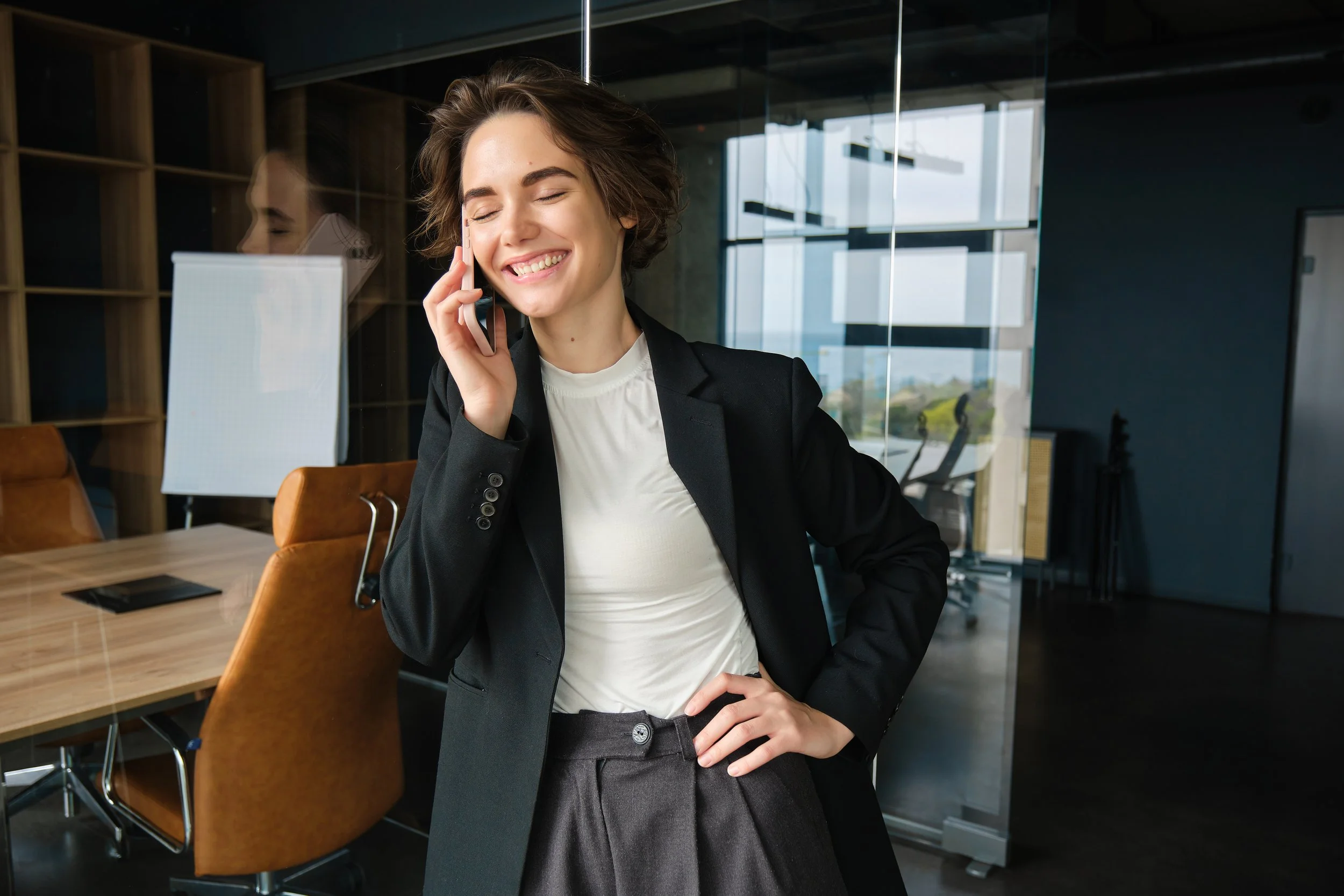 A young woman in business attire is talking on her phone in an office conference room, smiling and standing next to a large wooden table and office chairs, with large windows and modern decor in the background.
