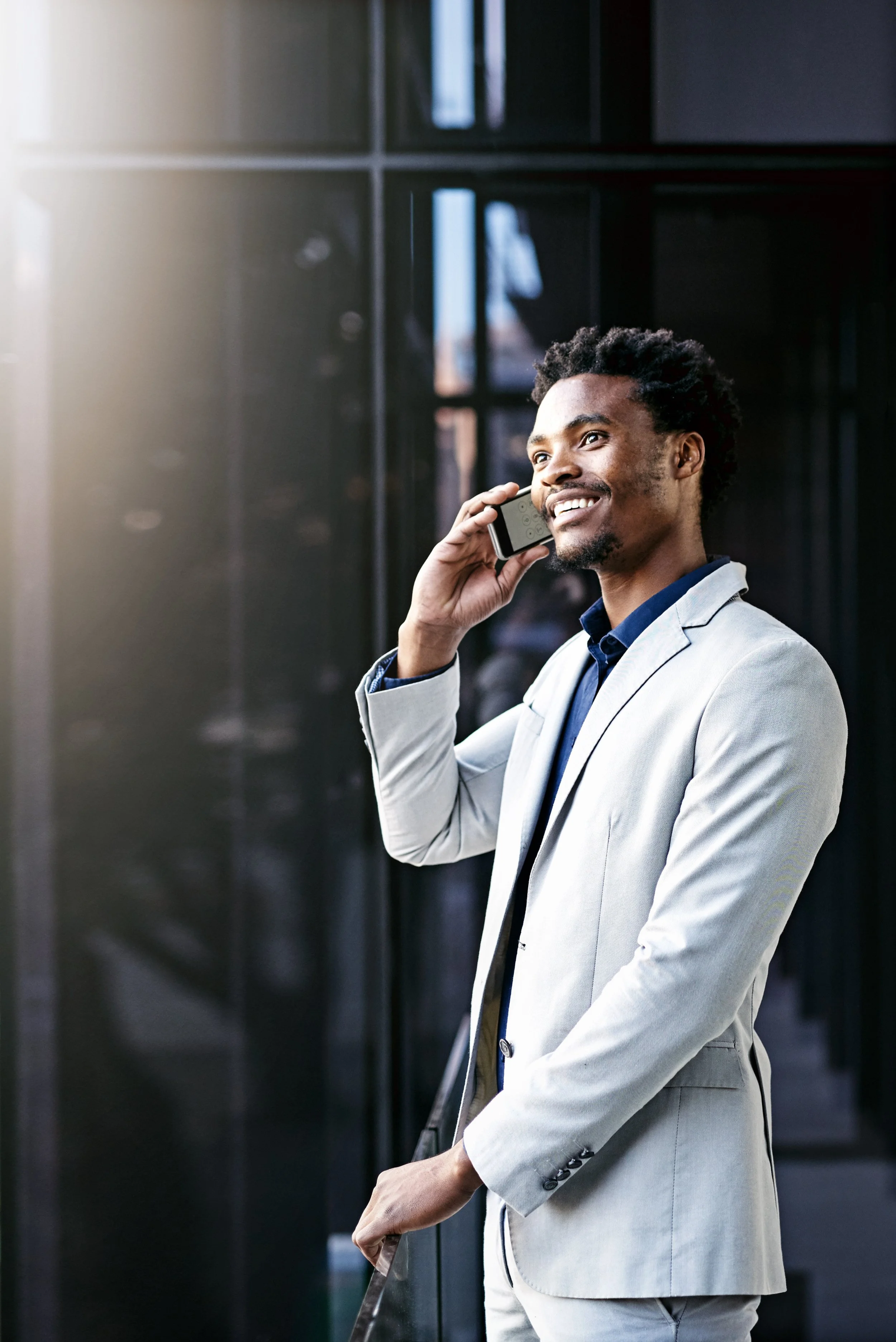 A young man in a light gray suit smiling and talking on his cell phone inside a modern building.