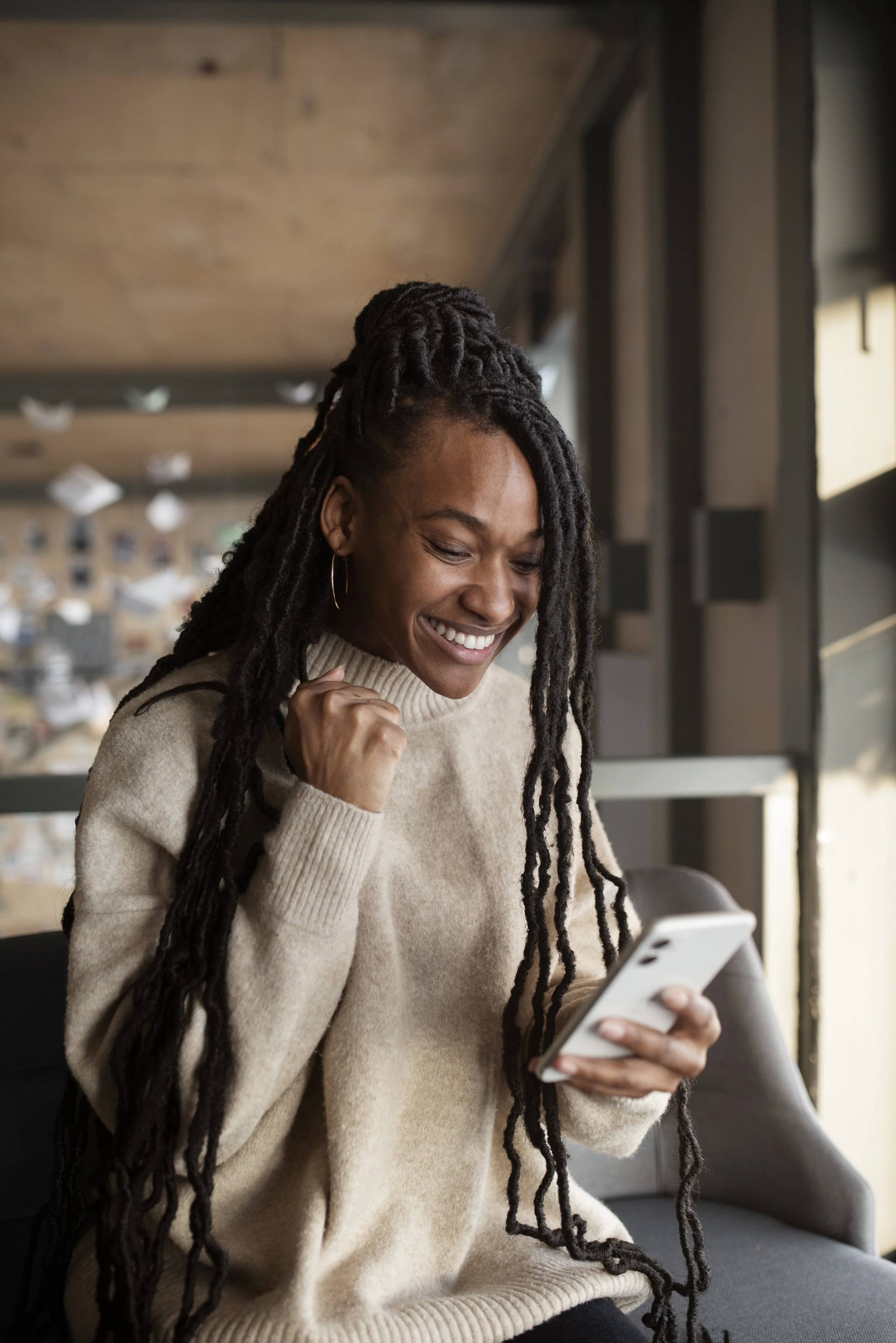 A young woman with dreadlocks, wearing a beige sweater, smiling and celebrating while looking at her smartphone in a cozy room with large windows.