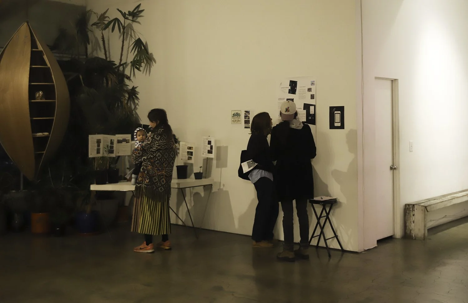 People in an art gallery at Navel in Los Angeles, viewing displays and engaging in conversations, with a large plant and a unique wooden display shelf in the background.