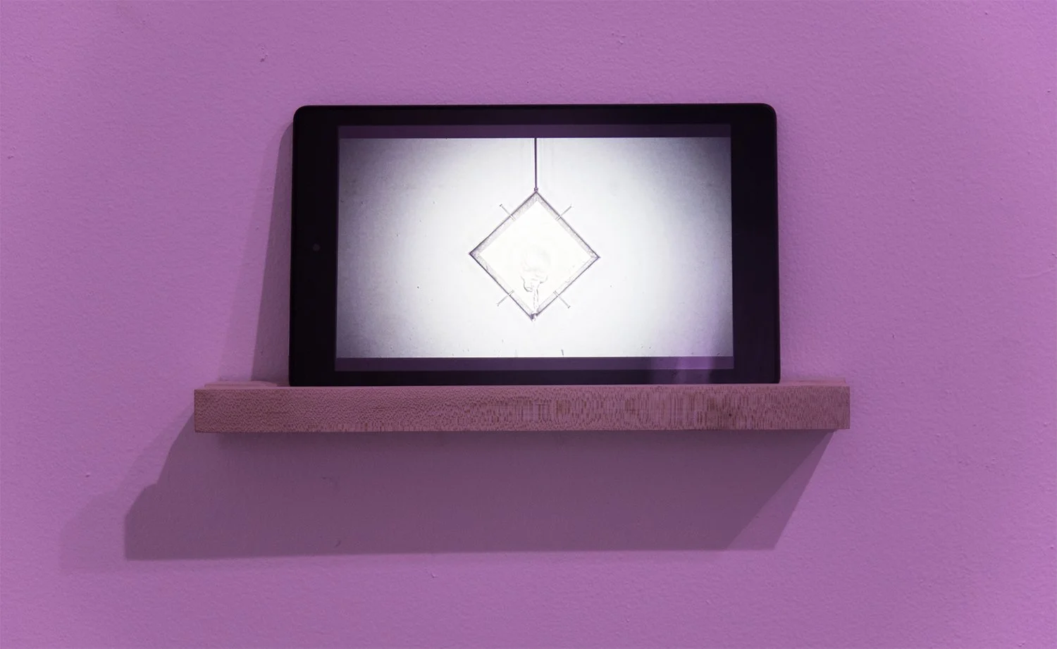 Digital artwork by Elisa Balmaceda on a tablet showing a geometric design with a diamond shape and pins, mounted on a pink wall with a wooden shelf.