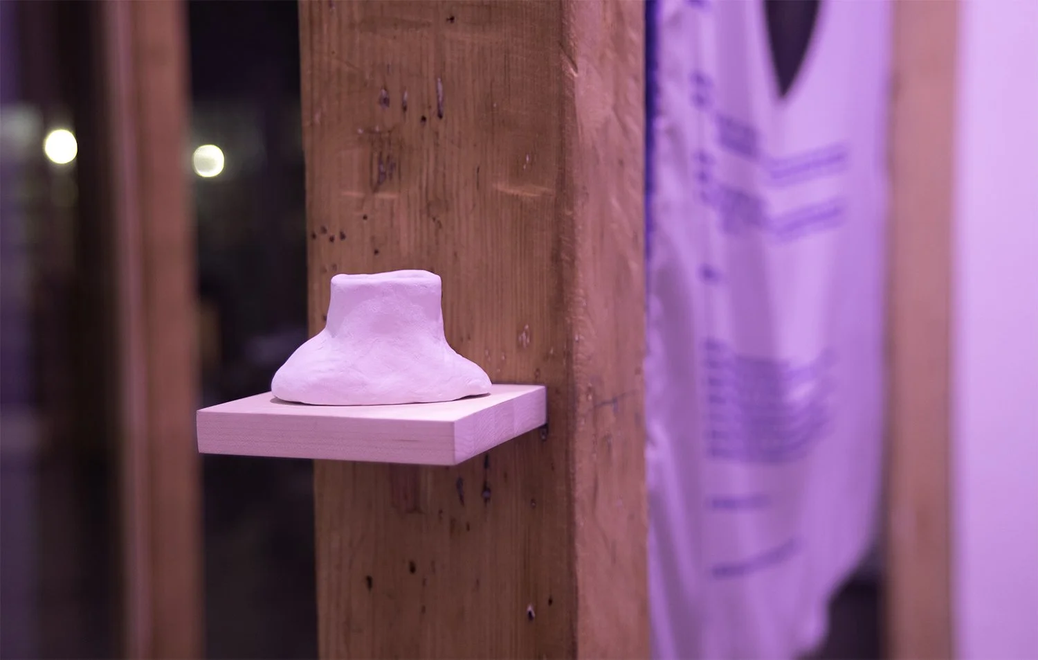 A irregularly shaped, pinkish ceramic volcano sculpture by Matteo Germain on a wooden shelf attached to a vertical wooden beam, under purple lighting.