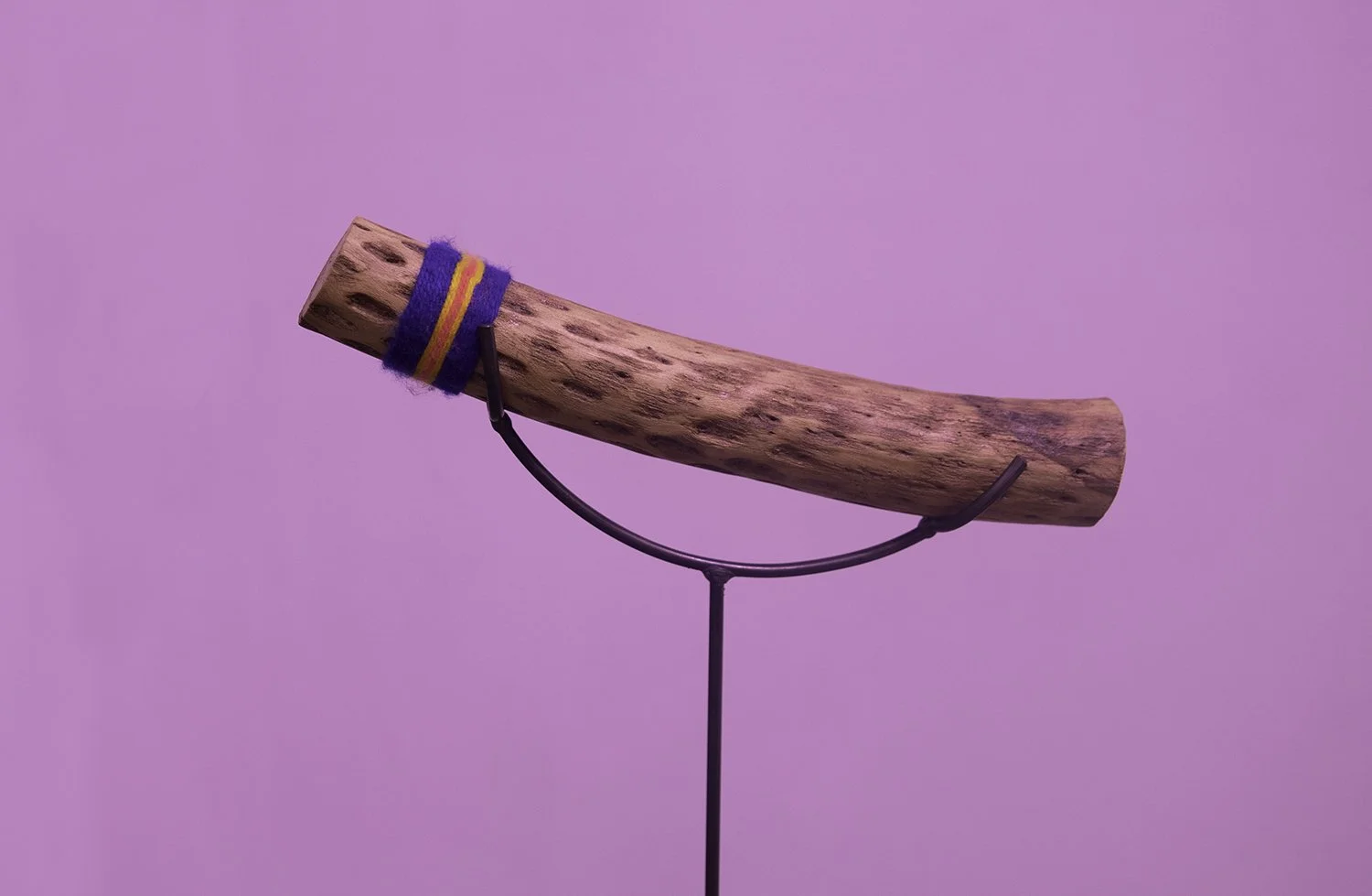 A wooden rain stick by Familia Villaroel, mounted on a black stand, with a colorful cloth wrapped around its end, against a pink background.