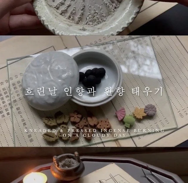 A collection of items on a table, including a white bowl with purple flowers, a plastic bag, decorative soaps shaped like animals and flowers, a lit candle, and some documents or books, created by artist Dambi Kim.