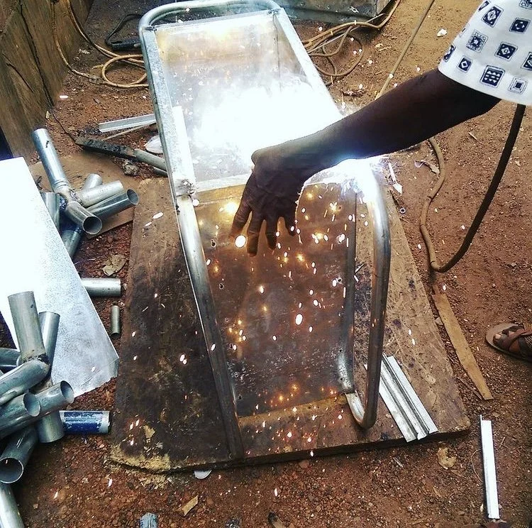 A person welding a metal chair outdoors, sparks flying from the welding process, with various metal pipes and tools scattered around, photo taken by architect Dk Osseo-Asare.