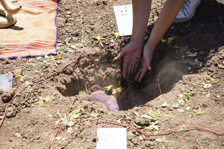 Artist Nicole L'Huillier is planting a vibrational device in a hole in the ground, with soil and gardening tools around.