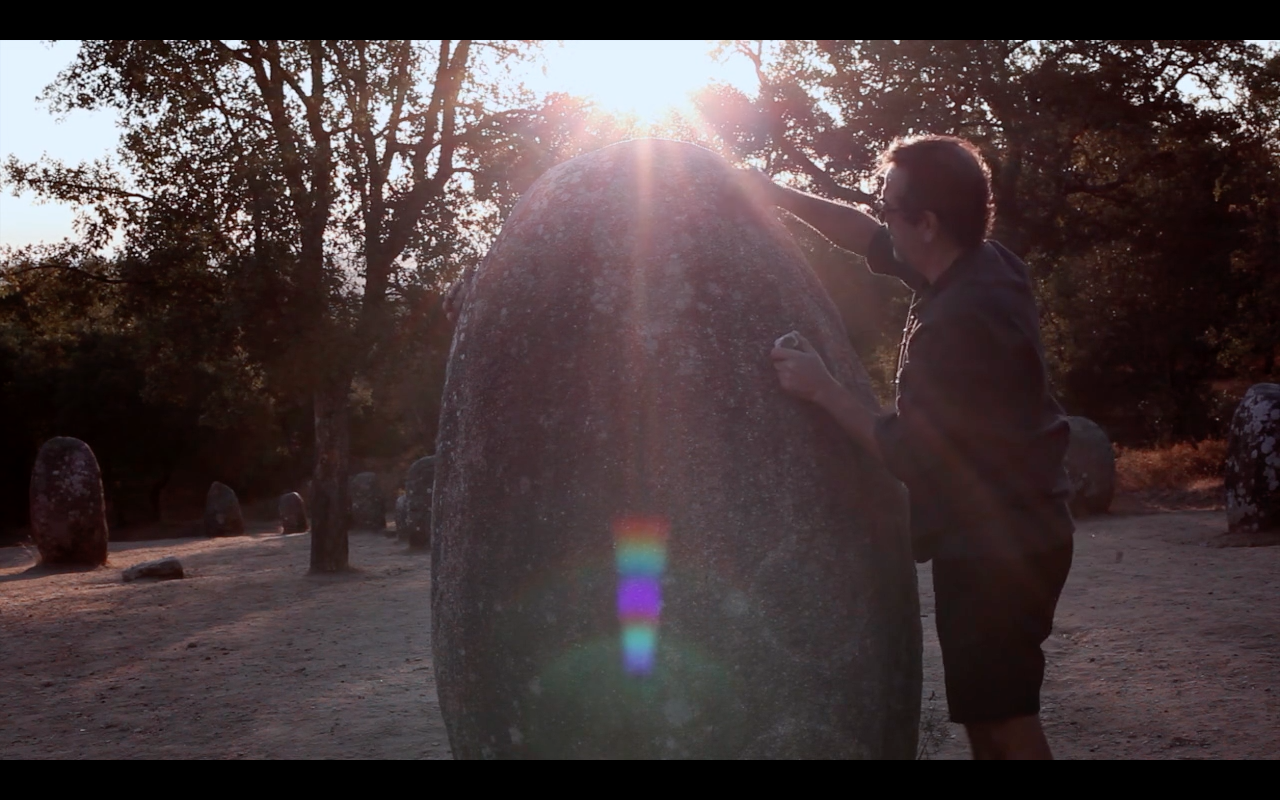 Man inspecting a large standing stone during sunset in a park with other stones and trees in the background, a film by artist Samuel Mountford.