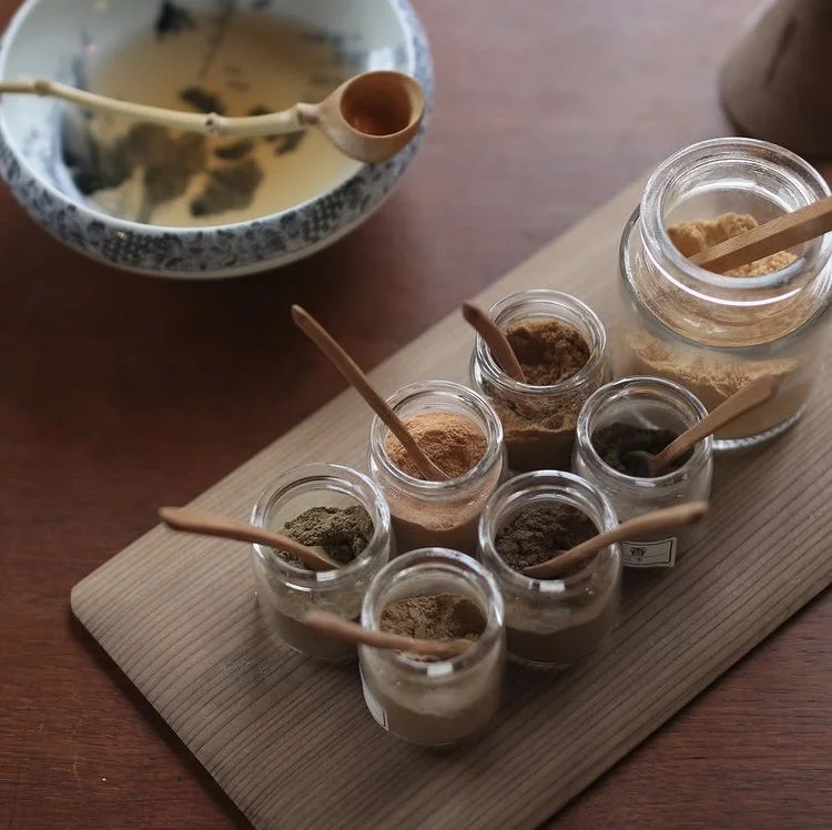 Assorted spices in small glass jars on a wooden board, with a tea pot in the background, set up by artist Dambi Kim.