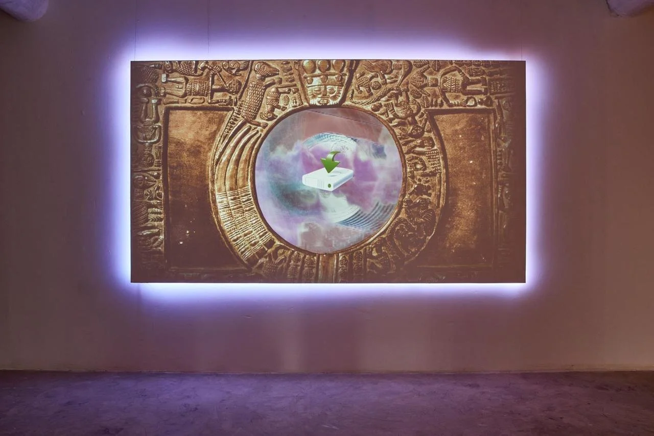 A wall-mounted artwork features an ancient coin or artifact with intricate carvings, illuminated by purple backlighting. A circular screen in the center displays a digital projection of a technological device, an artwork by Tabita Rezaire.