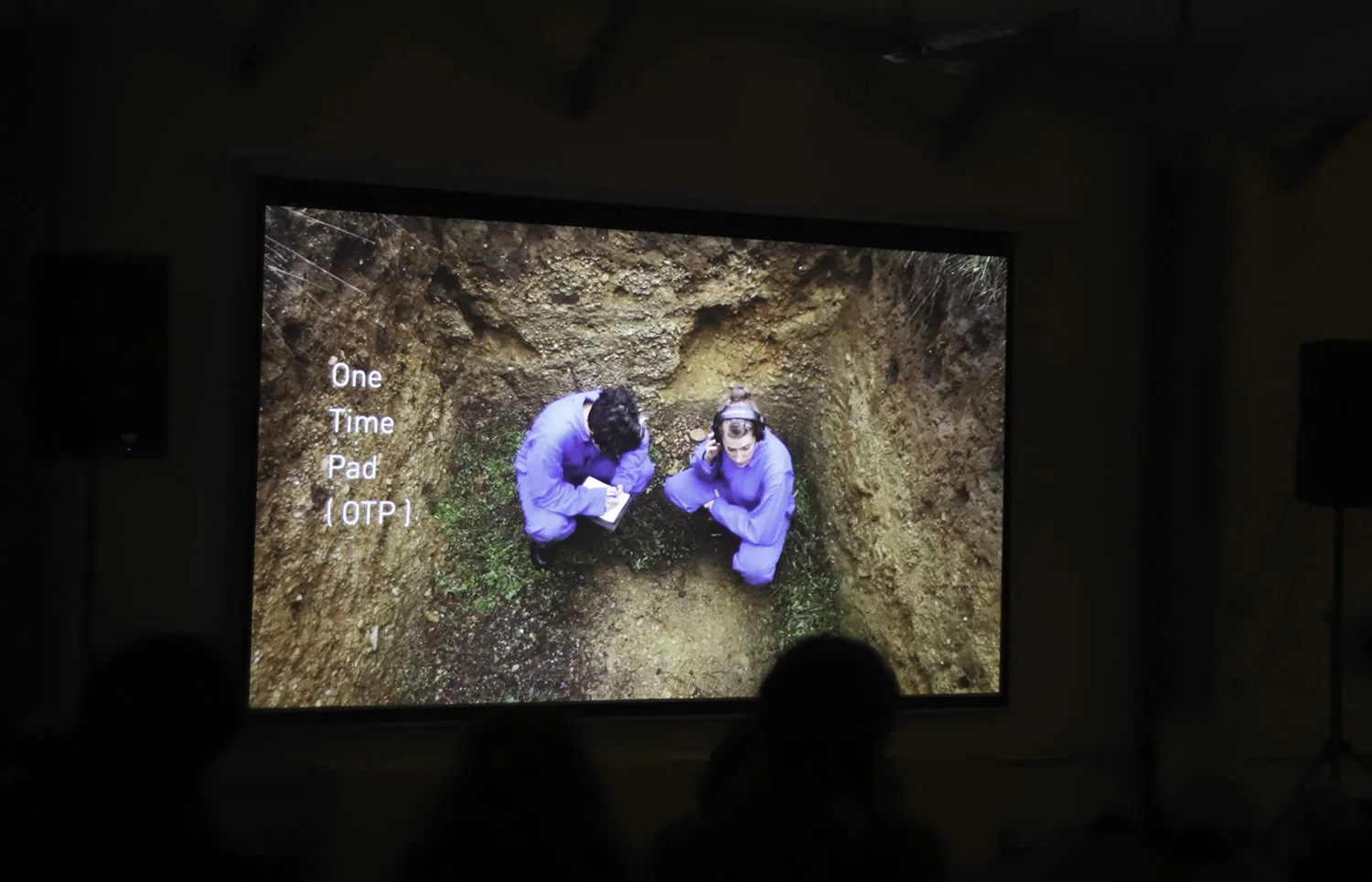 A presentation slide showing two people in blue outfits crouched inside a small excavated hole, with text reading 'One Time Pad (OTP)', a film by Nina Sarnelle and Sara Drake.