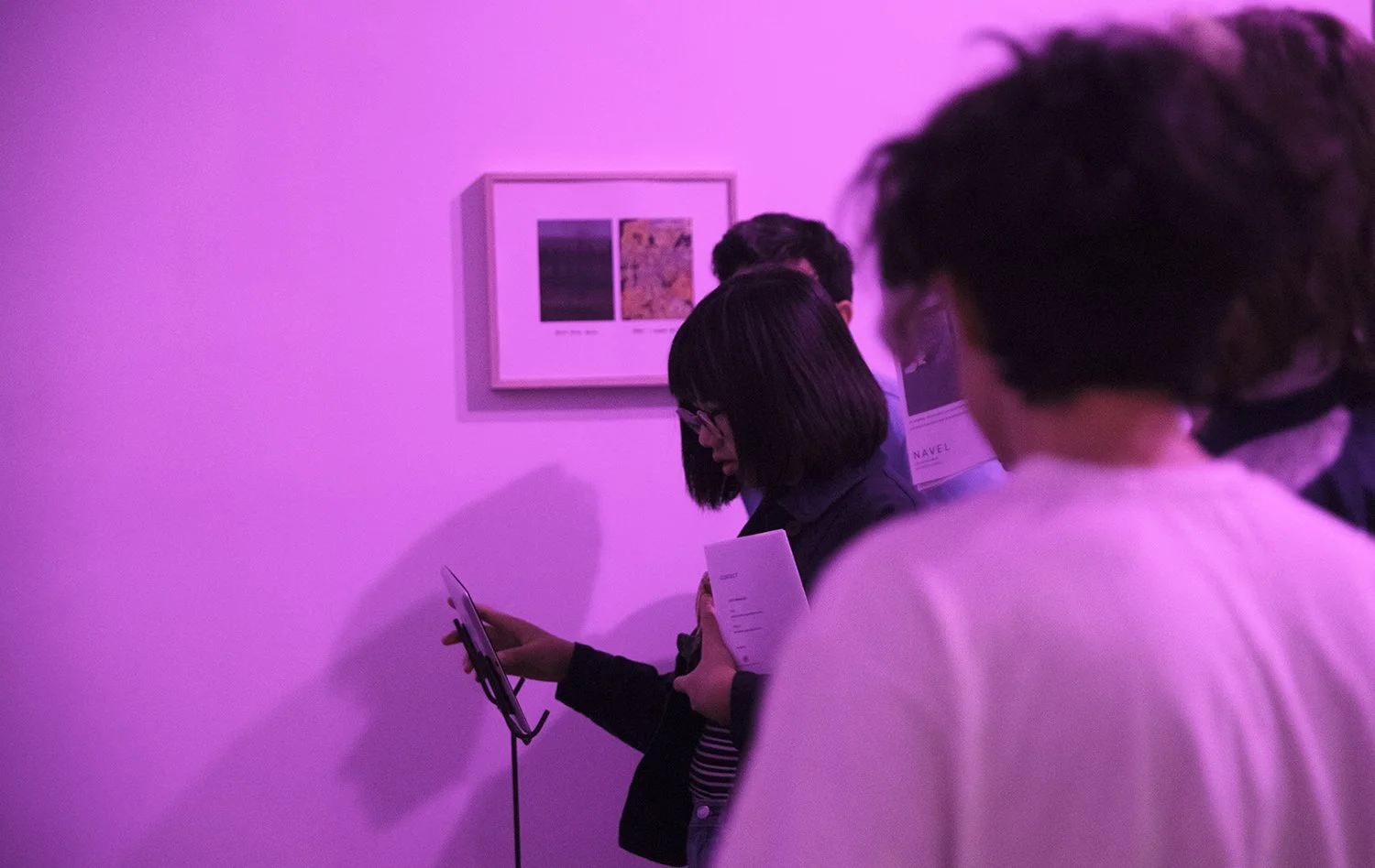 People standing in a purple-lit art gallery at Navel in Los Angeles, looking at artwork and reading brochures.