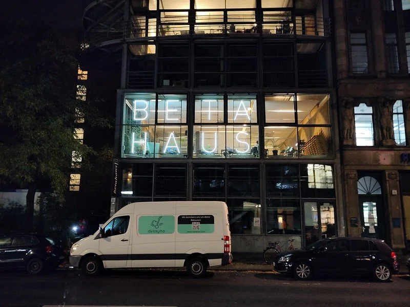 Glass building with illuminated neon sign spelling 'BETAHAUS' inside, parked white delivery van and cars on street at night in Berlin.