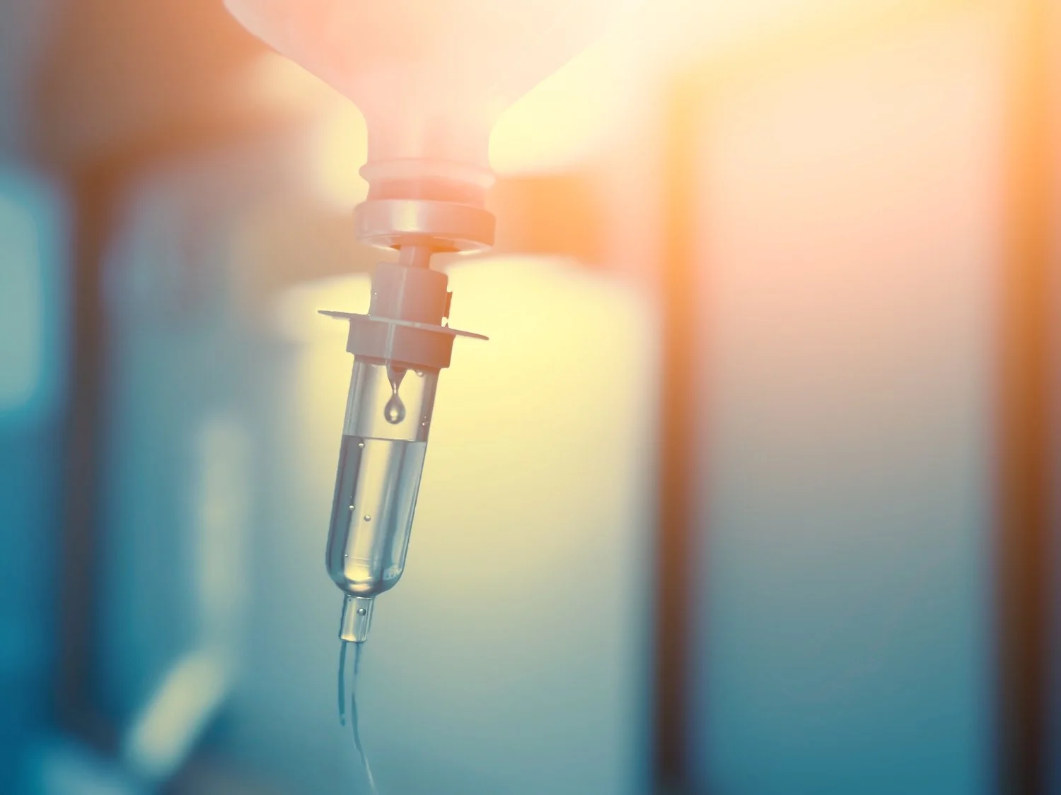 A close-up of a medical IV drip with a drop of fluid hanging from the needle, with soft lighting and blurred background.