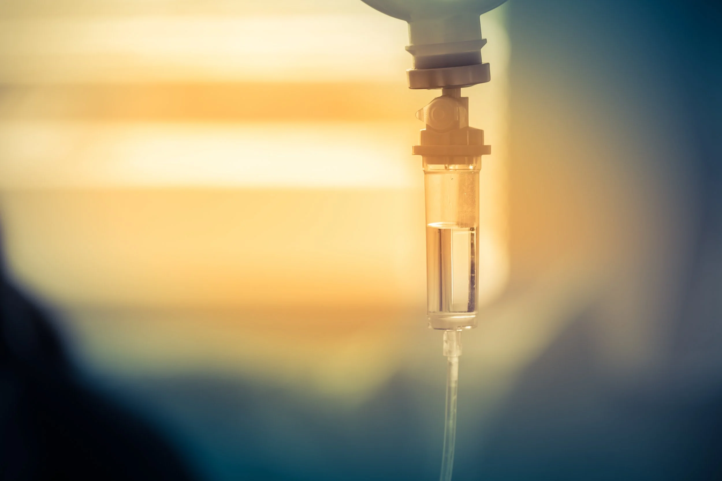 Close-up of an intravenous (IV) drip set with a fluid bag hanging, in a medical setting with warm lighting.