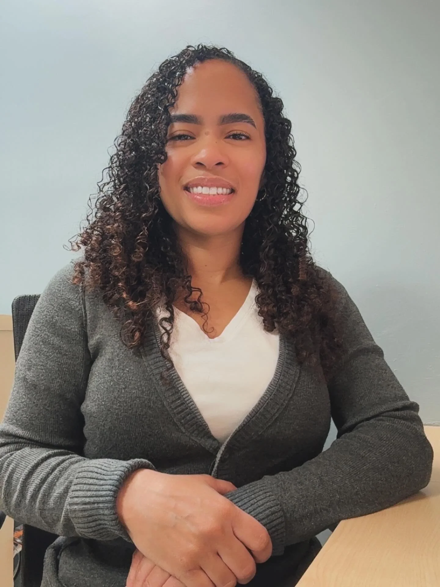 A woman with curly hair sitting at a desk, smiling, wearing a gray cardigan over a white shirt, in a professional setting.