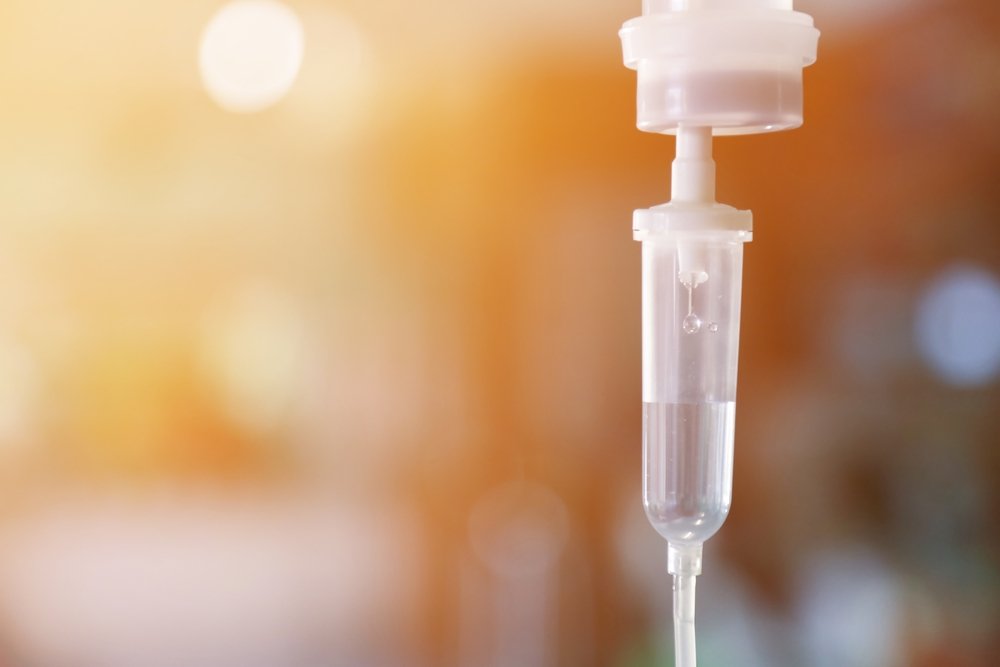 Close-up of a medical intravenous (IV) drip hanging, with a blurred warm background.