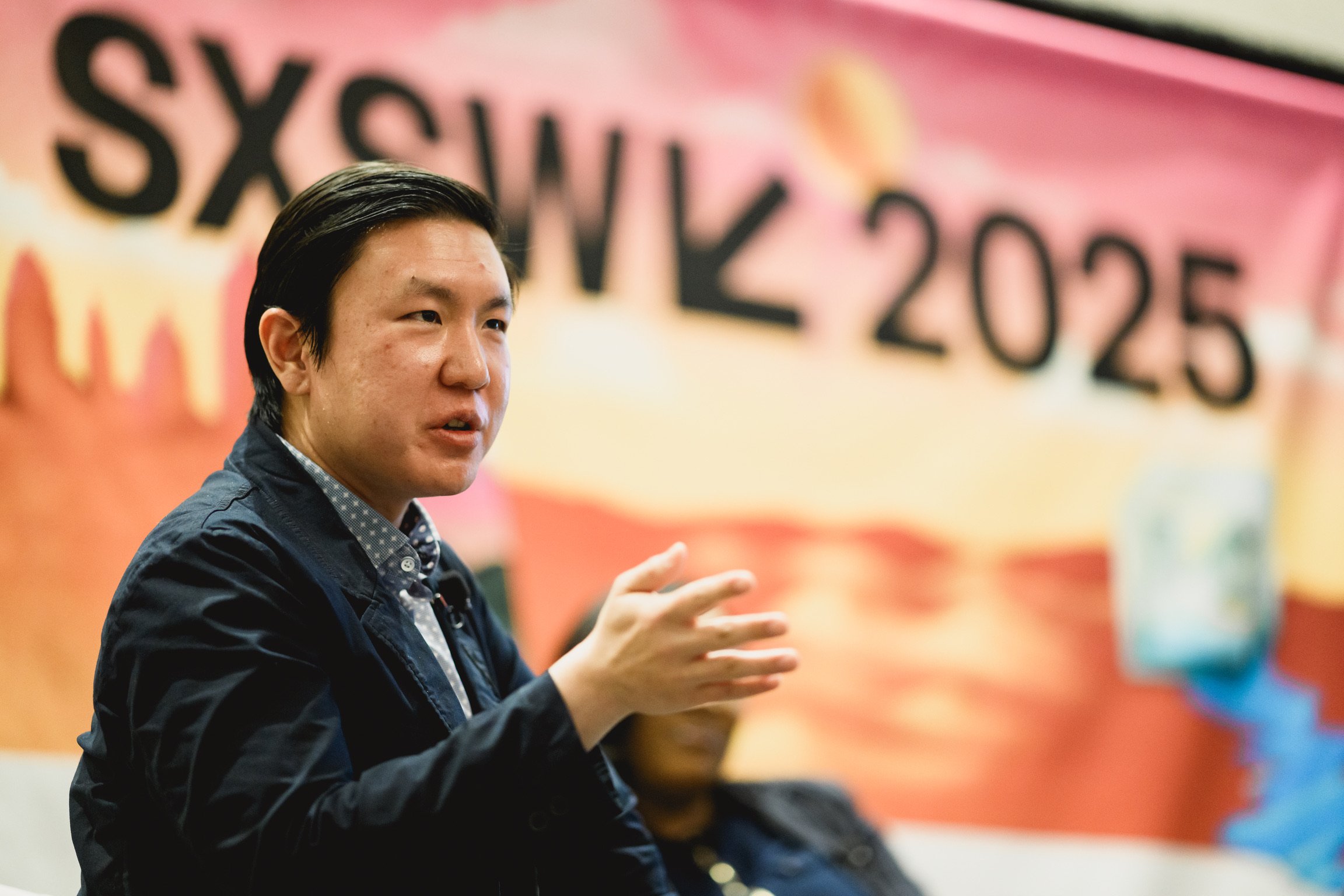 James Wang speaking or explaining, gesturing with his hand. Behind him is a colorful banner with the text 'SXSW 2025' in large black letters.