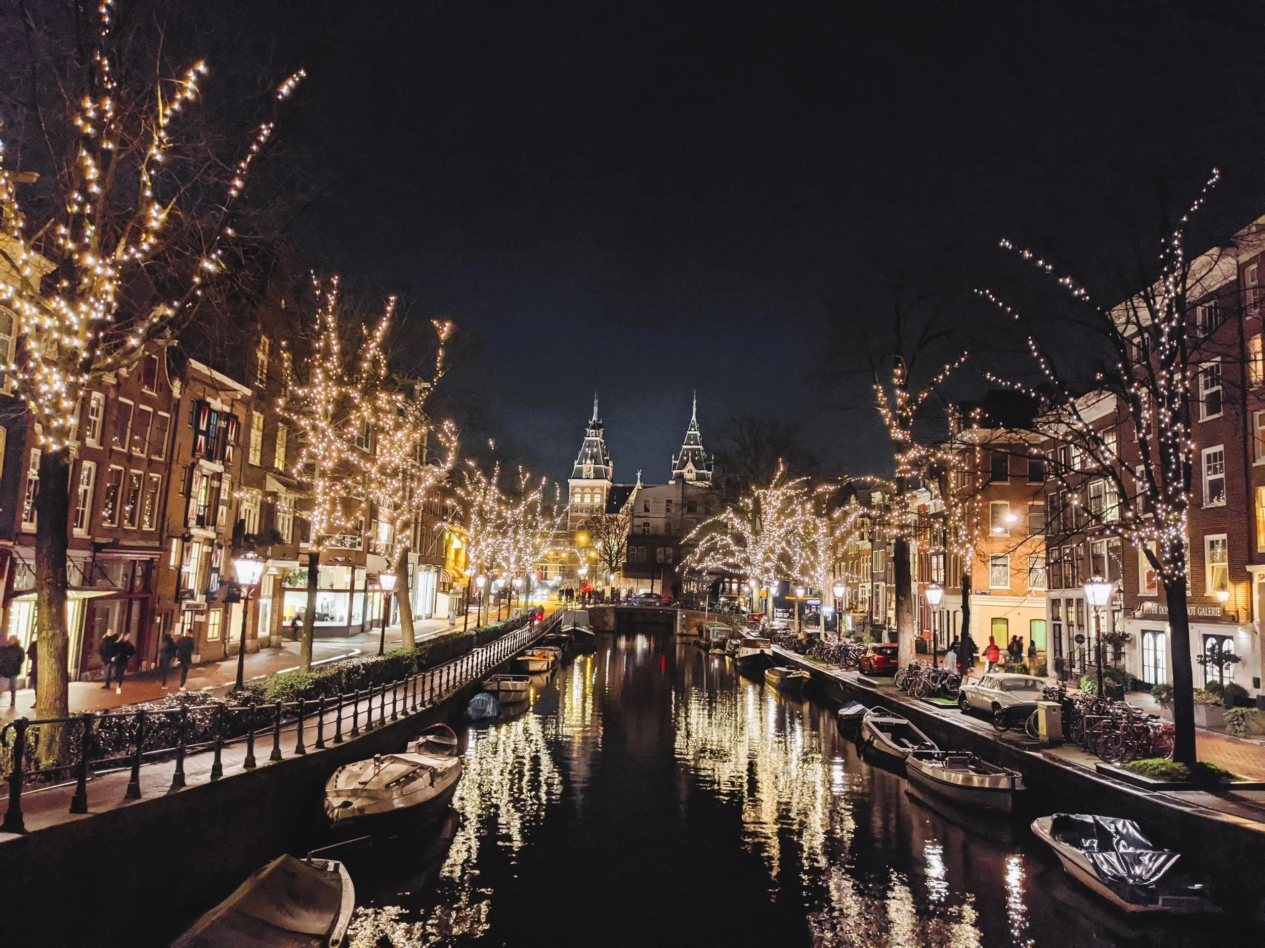 Night scene of a canal lined with buildings and illuminated trees decorated with string lights, with boats docked along the canal and a church with twin steeples in the background.