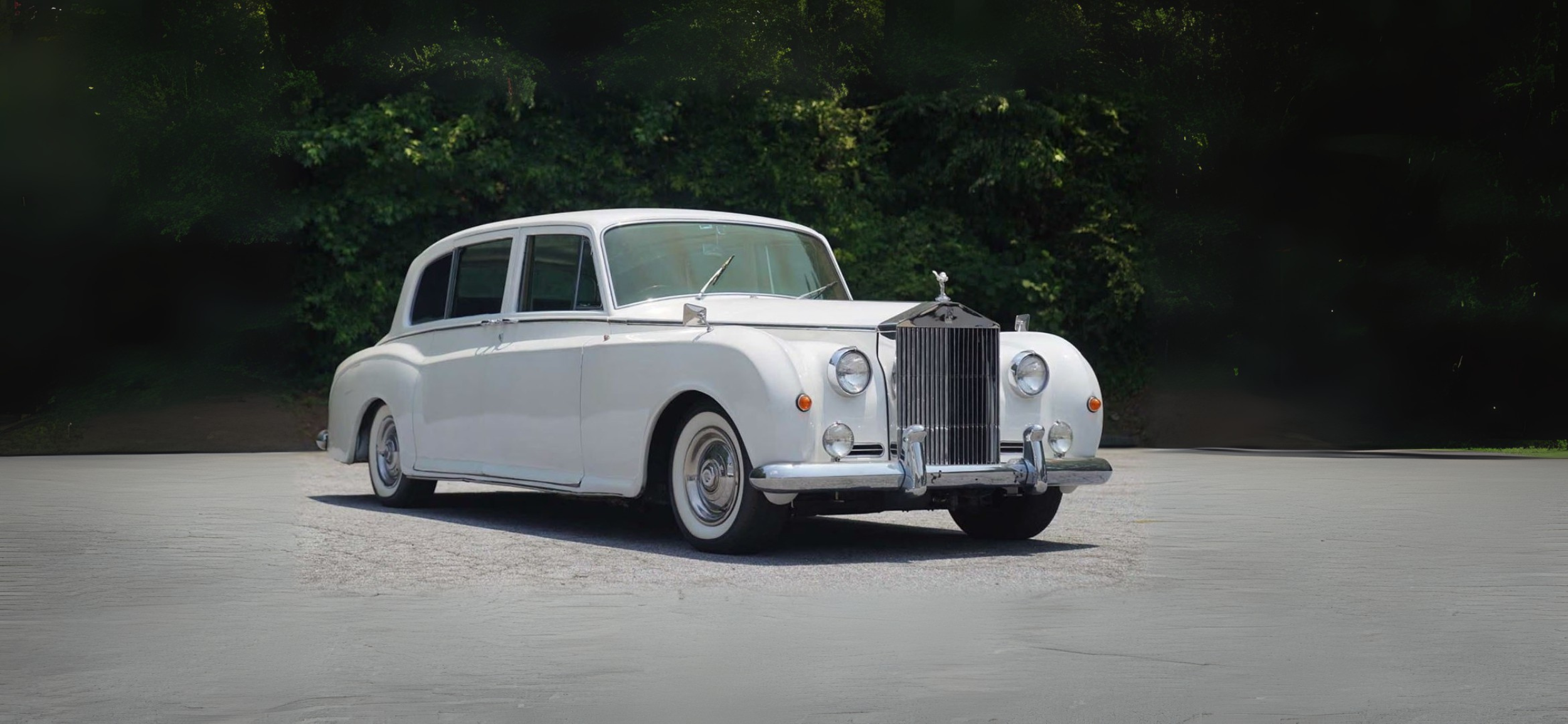 A vintage white luxury car parked on a paved area with a lush green forest in the background.